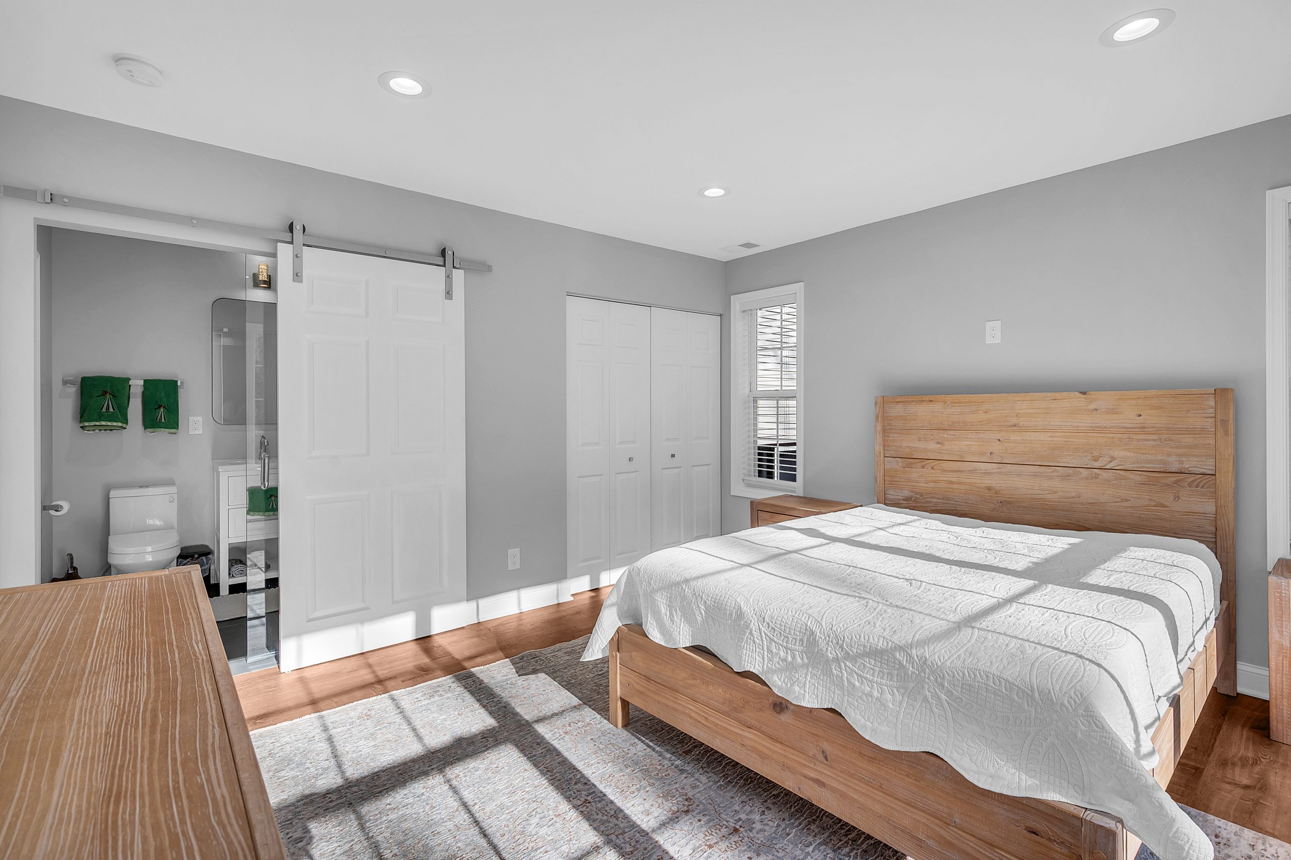 Bright and modern bedroom part of a room addition project in Fairfax, showcasing a wooden queen-sized bed with a crisp white quilt, polished hardwood floors, and a peek into the attached bathroom with green towels, all designed by Mosaic Design & Build