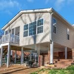 Rear view of a two-story house showcasing a new room addition on the first level, with a spacious deck above it, set against a backdrop of a well-kept lawn and clear blue sky